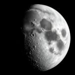 Superfície lunar acidentada em tons monocromáticos com uma cratera de impacto recente e poeira brilhante dispersa ao redor.